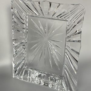 May include: A rectangular clear glass picture frame featuring a central starburst pattern etched into the glass. The frame has a wide, wavy border with deep, vertical cut grooves that create a textured, light-reflecting surface around the edges.