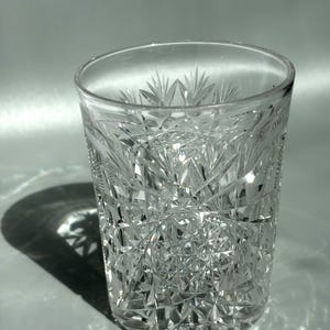 Könnte beinhalten: Ein klarer Trinkbecher mit einem komplexen Schliffmuster aus sich kreuzenden Linien, Rauten und Sternmotiven. Das Gefäß mit schwerem Boden reflektiert das Licht und erzeugt scharfe geometrische Schatten und Glanzlichter auf der facettierten Oberfläche.