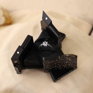 Puede incluir: Un joyero negro y geométrico con un anillo de compromiso plateado en su interior. El anillo tiene una piedra central grande y transparente y piedras más pequeñas en la banda. La caja está abierta, revelando el anillo contra un interior de terciopelo negro.