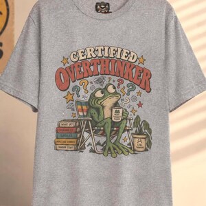 May include: A heather grey t-shirt featuring a graphic of a green frog sitting in a folding chair, holding a mug that says "MORE THOUGHTS PLEASE". The text "CERTIFIED OVERTHINKER" is arched above the frog, with a stack of books and a potted plant.