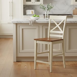 May include: A cream-colored wooden bar stool with a brown seat and an X-shaped backrest. The stool is positioned in front of a kitchen island with a marble countertop. The kitchen has light-colored cabinets and hardwood floors.