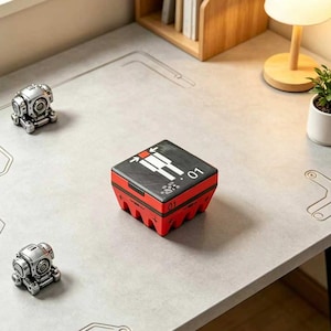 May include: A red and black square robot with a graphic on top and the number "01" is displayed on a gray surface. Two small silver robots are also on the surface. The robots appear to be toys or models.