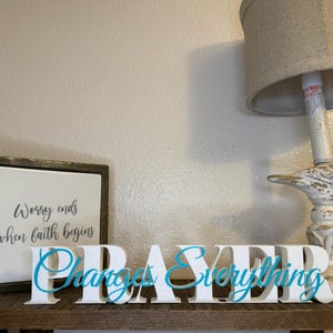 May include: A square sign with a dark wood frame and white center featuring the black text 'Worry ends when faith begins' next to a white block letter sign spelling 'PRAYER' with the blue script text 'Changes Everything' layered over it.
