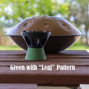 May include: A black and green stand with a leaf pattern, positioned in front of a large, metallic, dome-shaped handpan instrument. The stand features a series of circular cutouts around its upper section and a textured green base.