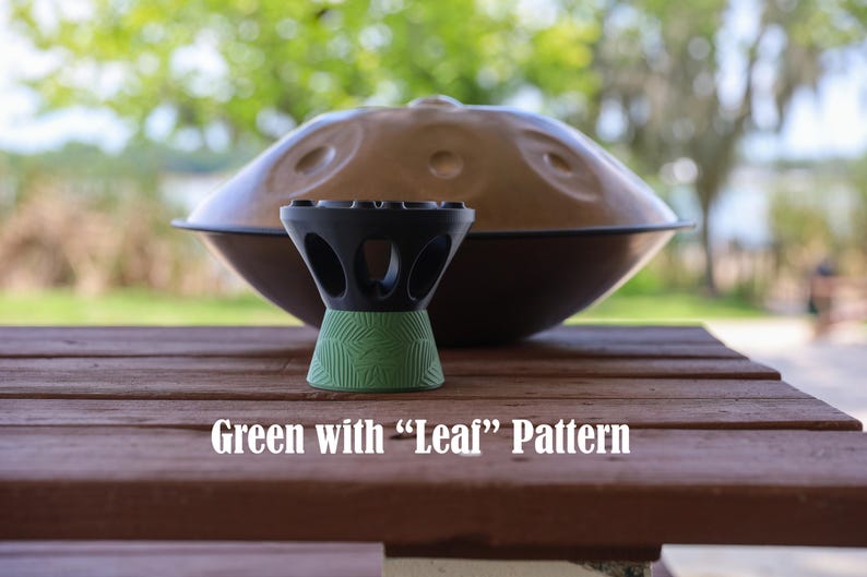 May include: A black and green stand with a leaf pattern, positioned in front of a large, metallic, dome-shaped handpan instrument. The stand features a series of circular cutouts around its upper section and a textured green base.