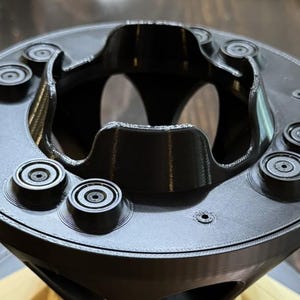 May include: A black, circular, 3D-printed component featuring a central opening with curved, scalloped edges and a flat top surface. Six small, raised, circular protrusions are evenly spaced around the perimeter of the top face.