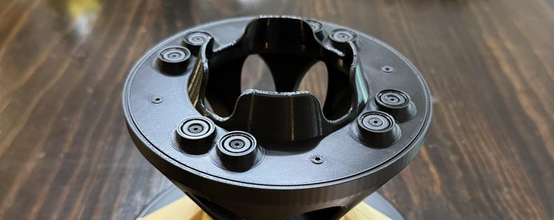 May include: A black, circular, 3D-printed component featuring a central opening with curved, scalloped edges and a flat top surface. Six small, raised, circular protrusions are evenly spaced around the perimeter of the top face.