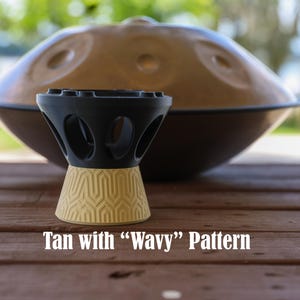 May include: A black and tan stand for a handpan instrument. The top section is black with circular cutouts, while the base is tan with a textured wavy geometric pattern. The text "Tan with "Wavy" Pattern" is printed below the stand.