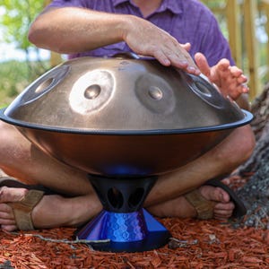 May include: A metallic bronze handpan drum with a series of circular tone fields on its upper surface. The instrument is supported by a blue, multi-faceted, geometric stand featuring a hollowed-out design with several oval-shaped openings.