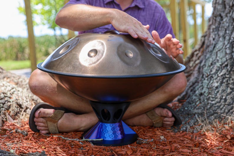 May include: A metallic bronze handpan drum with a series of circular tone fields on its upper surface. The instrument is supported by a blue, multi-faceted, geometric stand featuring a hollowed-out design with several oval-shaped openings.