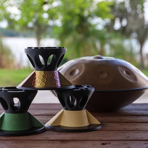 May include: Three black stands with colorful bases are arranged in a pyramid formation. The top stand has a metallic honeycomb pattern, the bottom left has a green chevron design, and the bottom right has a tan geometric pattern. A brown handpan sits behind.