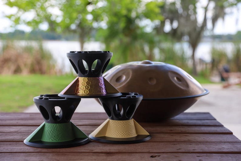 May include: Three black stands with colorful bases are arranged in a pyramid formation. The top stand has a metallic honeycomb pattern, the bottom left has a green chevron design, and the bottom right has a tan geometric pattern. A brown handpan sits behind.