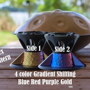 May include: Two black stands with a hex pattern and a 4 color gradient shifting blue, red, purple, and gold finish. The stands are labeled Side 1 and Side 2. A large brown handpan instrument is positioned in the background behind the stands.