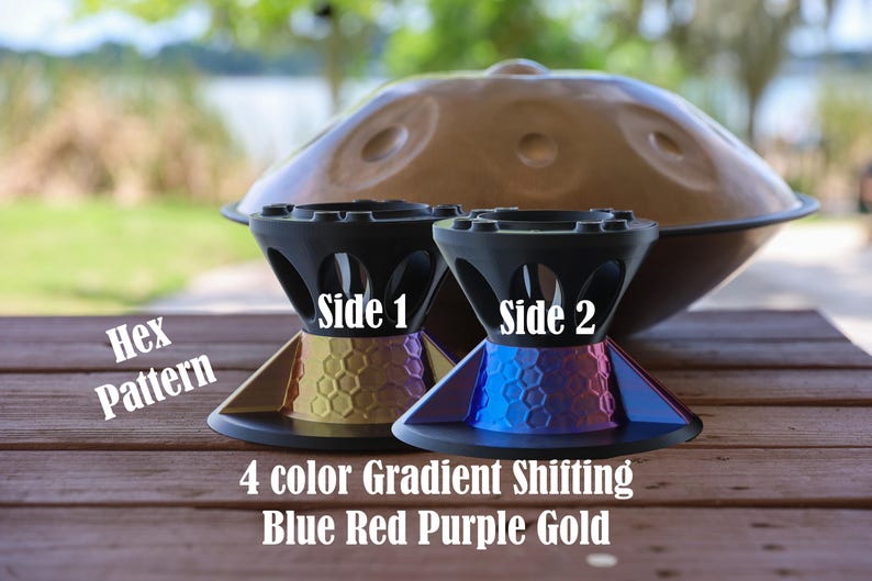 May include: Two black stands with a hex pattern and a 4 color gradient shifting blue, red, purple, and gold finish. The stands are labeled Side 1 and Side 2. A large brown handpan instrument is positioned in the background behind the stands.