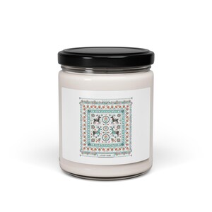 Pode incluir: Um frasco de vela branco com tampa preta. O rótulo apresenta um design quadrado com veados, flores e padrões geométricos em verde-azulado, vermelho e branco. O texto "HYGGE HOME" está na parte inferior.