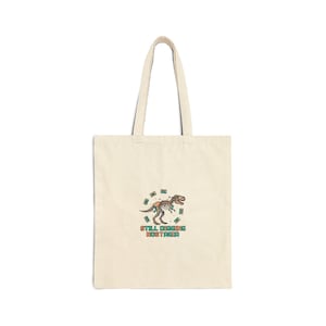 May include: Cream-colored tote bag with a dinosaur skeleton graphic and the text "Still Digging Nostalgia." The graphic features a T-Rex skeleton with green and brown accents, surrounded by green dollar bill illustrations.