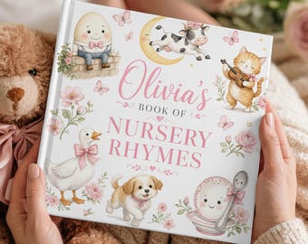 May include: A hardcover children's book titled "Olivia's Book of Nursery Rhymes" with illustrations of a cow on a crescent moon, a cat playing a violin, and other cute characters. The book is held by a person, next to a teddy bear.