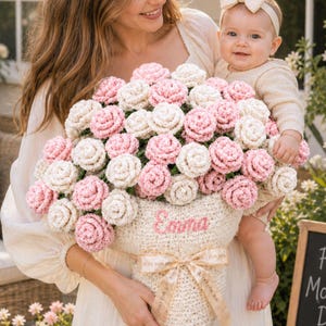 Puede incluir: Una mujer y un bebé sostienen una gran cesta de rosas rosas y blancas tejidas a ganchillo. La cesta es de color crema y tiene bordado el nombre "Emma". Un letrero de pizarra dice "First Mother's Day".