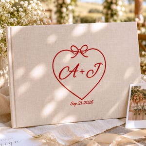 Puede incluir: Un libro de visitas rectangular con cubierta de lino beige que presenta un bordado rojo de un corazón con un lazo, las letras A+J en el centro y la fecha Sep. 21, 2026, bajo el corazón, acompañado de una pequeña fotografía de cuatro personas.