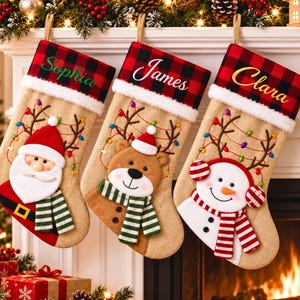 Può includere: Tre calze natalizie in juta con risvolti a quadri rossi e neri, personalizzate con i nomi ricamati Sophia, James e Clara. Le calze presentano applicazioni in peluche di Babbo Natale, un orso e un pupazzo di neve con luci decorative.
