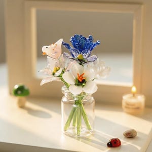 Puede incluir: Un pequeño jarrón de cristal contiene un ramo de flores de cristal en tonos blancos, azules y rosas. También se ven un pequeño pájaro de cristal y una mariquita. El arreglo está sobre una superficie blanca.