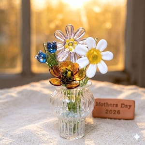 May include: A clear glass vase holds a colorful bouquet of glass flowers. The flowers include white daisies, orange and yellow blooms, and blue buds. The vase sits on a white surface, with a brick that reads "Mothers Day 2026".