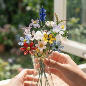 May include: A colorful glass flower bouquet in a clear vase. The flowers are various colors, including red, blue, yellow, and white. The bouquet is held in a clear glass vase. The background is blurred, with green foliage visible.