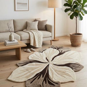 May include: A large, irregularly shaped area rug featuring a stylized floral design in shades of cream, beige, and dark brown. The rug has a tufted texture with curved, petal-like sections radiating from the center, creating a distinct organic shape.