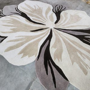 May include: A large, irregularly shaped area rug featuring a stylized floral design. The rug is composed of cream, beige, taupe, and dark brown tufted sections that create a layered petal effect, radiating from a central point in a flower pattern.