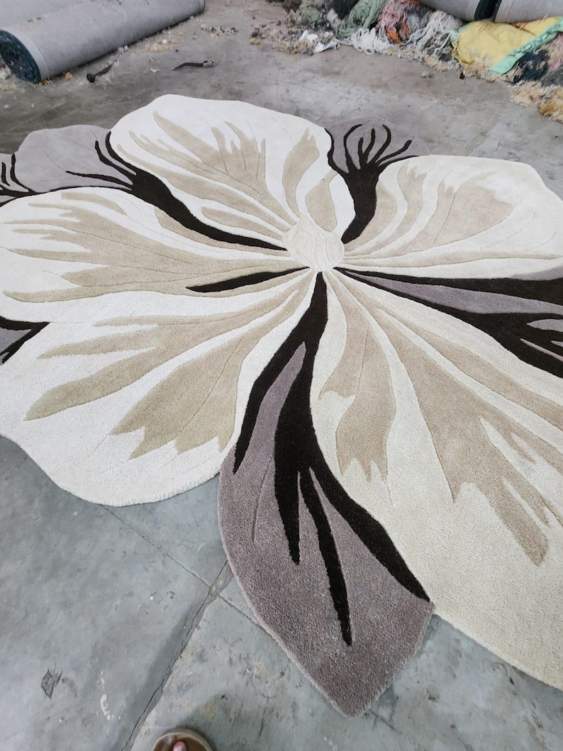 May include: A large, irregularly shaped area rug featuring a stylized floral design. The rug is composed of cream, beige, taupe, and dark brown tufted sections that create a layered petal effect, radiating from a central point in a flower pattern.
