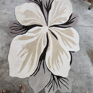 May include: A large, flower-shaped area rug featuring a layered petal design in shades of cream, beige, and dark brown. The rug has a soft, tufted texture with distinct, dark brown vein-like patterns radiating from the center toward the edges.