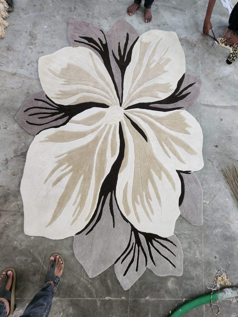May include: A large, flower-shaped area rug featuring a layered petal design in shades of cream, beige, and dark brown. The rug has a soft, tufted texture with distinct, dark brown vein-like patterns radiating from the center toward the edges.