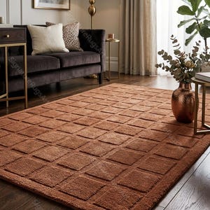 May include: A terracotta-coloured rectangular rug featuring a repeating square design. The rug is laid on a dark wooden floor in a living room, with a dark brown sofa and gold accents in the background. The rug measures approximately 200cm x 300cm.