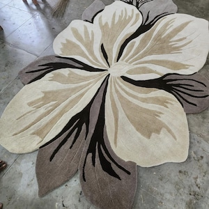 May include: A large, flower-shaped area rug featuring a layered petal design in shades of cream, beige, taupe, and dark brown. The rug has a tufted texture with distinct, curved lines radiating from the center, mimicking the organic form of a blossom.