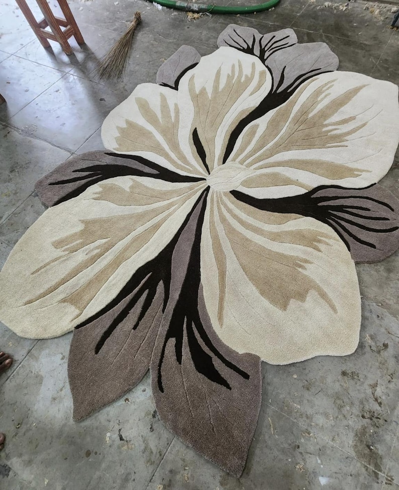 May include: A large, flower-shaped area rug featuring a layered petal design in shades of cream, beige, taupe, and dark brown. The rug has a tufted texture with distinct, curved lines radiating from the center, mimicking the organic form of a blossom.