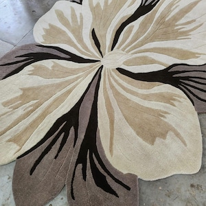May include: A large, flower-shaped area rug featuring a layered petal design in shades of cream, beige, taupe, and dark brown. The rug has a textured, tufted surface with distinct color blocking that defines the organic, curved shapes of the petals.