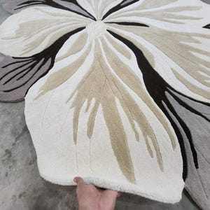 May include: A large, flower-shaped area rug featuring a tufted texture. The design consists of organic, petal-like segments in shades of cream, beige, dark brown, and gray, radiating from a central point to create a bold, oversized floral pattern.