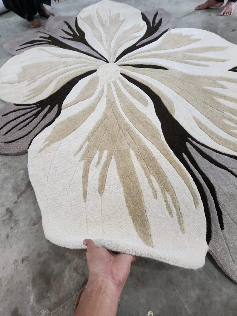May include: A large, flower-shaped area rug featuring a tufted texture. The design consists of organic, petal-like segments in shades of cream, beige, dark brown, and gray, radiating from a central point to create a bold, oversized floral pattern.