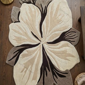 May include: A tufted area rug shaped like a large flower with organic, irregular edges. The design features a color palette of cream, beige, and dark brown, with deep brown accents radiating from the center to define the petal-like segments.