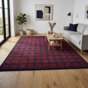 Puede incluir: Una alfombra rectangular grande con un patrón de cuadrícula geométrica en tonos rojo y azul marino. La alfombra está colocada sobre un suelo de madera en un salón, con un sofá, una mesa de centro y obras de arte visibles en el fondo.