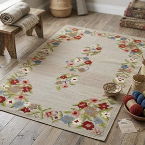 Op de afbeelding: Een rechthoekig beige tapijt met een bloemenrand in rood, blauw en groen. Het tapijt heeft een centraal bloemmotief. Rotan manden, garen en een kleine houten kruk omringen het tapijt. Het tapijt ligt op een houten vloer.
