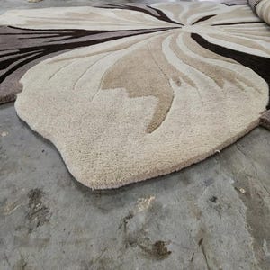 May include: A large, plush area rug featuring a floral-inspired pattern with organic, curved shapes. The rug is composed of varying shades of cream, beige, tan, and dark brown, with a thick, tufted texture that creates depth and dimension.