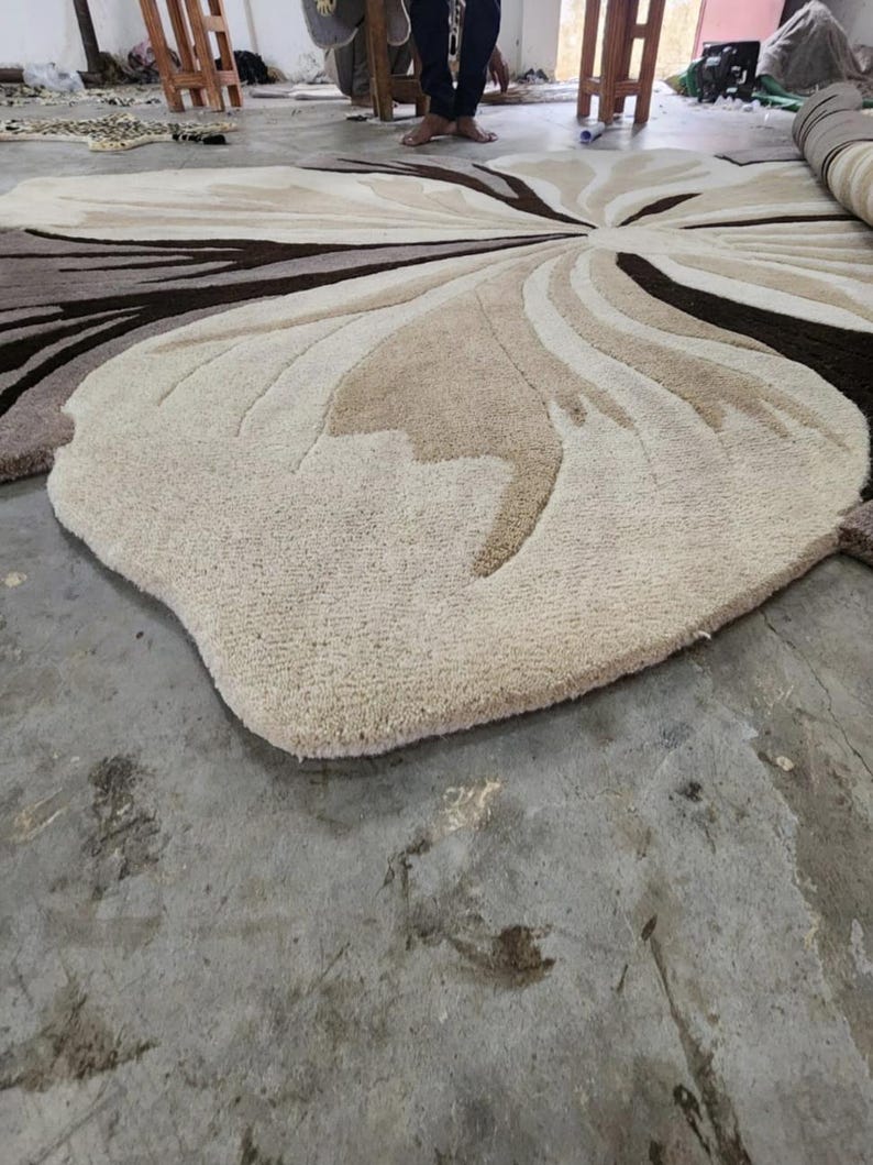 May include: A large, plush area rug featuring a floral-inspired pattern with organic, curved shapes. The rug is composed of varying shades of cream, beige, tan, and dark brown, with a thick, tufted texture that creates depth and dimension.