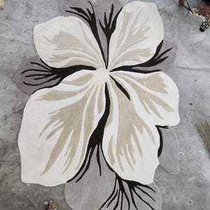May include: A large, irregularly shaped area rug featuring a stylized floral design. The rug is composed of cream, beige, and dark brown sections with deep black accents that define the petal shapes and create a high-contrast, layered appearance.