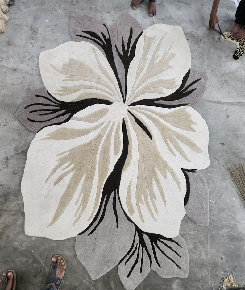 May include: A large, irregularly shaped area rug featuring a stylized floral design. The rug is composed of cream, beige, and dark brown sections with deep black accents that define the petal shapes and create a high-contrast, layered appearance.