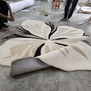 May include: A large, flower-shaped area rug featuring a tufted design in shades of cream, beige, and dark brown. The rug has a soft, textured surface with organic, petal-like contours and a curved edge, showcasing a layered, multi-toned floral pattern.
