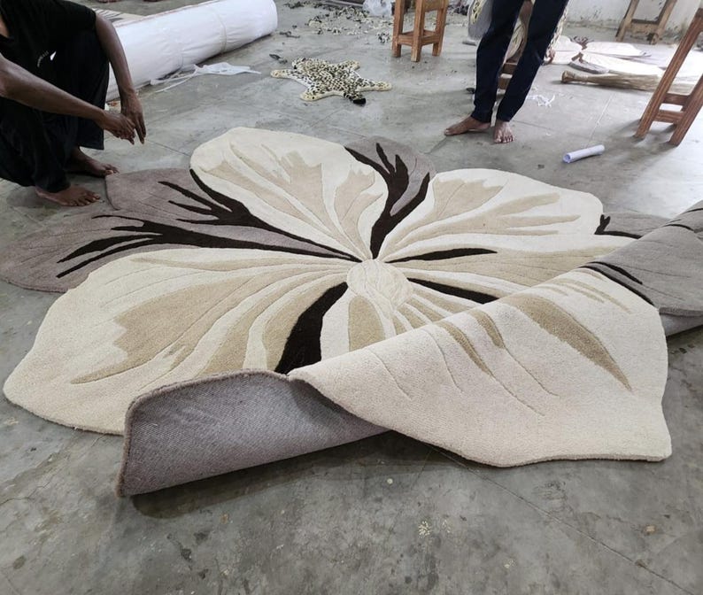 May include: A large, flower-shaped area rug featuring a tufted design in shades of cream, beige, and dark brown. The rug has a soft, textured surface with organic, petal-like contours and a curved edge, showcasing a layered, multi-toned floral pattern.