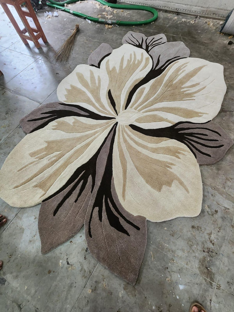 May include: A large, flower-shaped area rug featuring a layered petal design in shades of cream, beige, taupe, and dark brown. The rug has a tufted texture with distinct color blocking that defines the organic, curved edges of the floral motif.