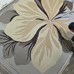 May include: A large rectangular rug in progress featuring a central oversized flower design. The floral motif is rendered in shades of cream, beige, and dark brown, set against a light gray background. The tufted texture is visible on the frame.