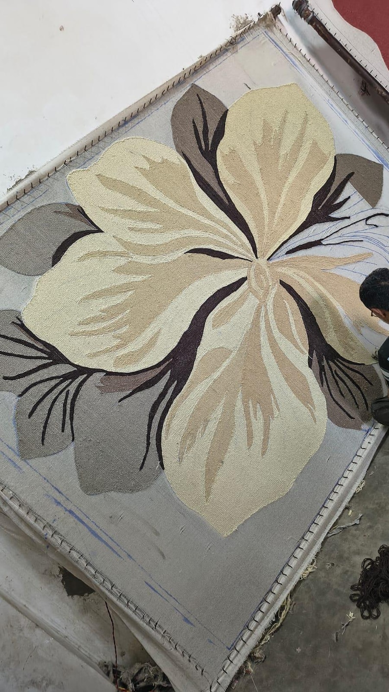 May include: A large rectangular rug in progress featuring a central oversized flower design. The floral motif is rendered in shades of cream, beige, and dark brown, set against a light gray background. The tufted texture is visible on the frame.
